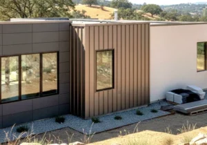 Maison en matériaux résistants entourée de végétation sèche, avec fumée lointaine visible